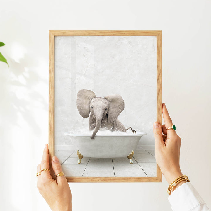 A person's hands are holding a framed picture of a baby elephant sitting in a bathtub filled with water.