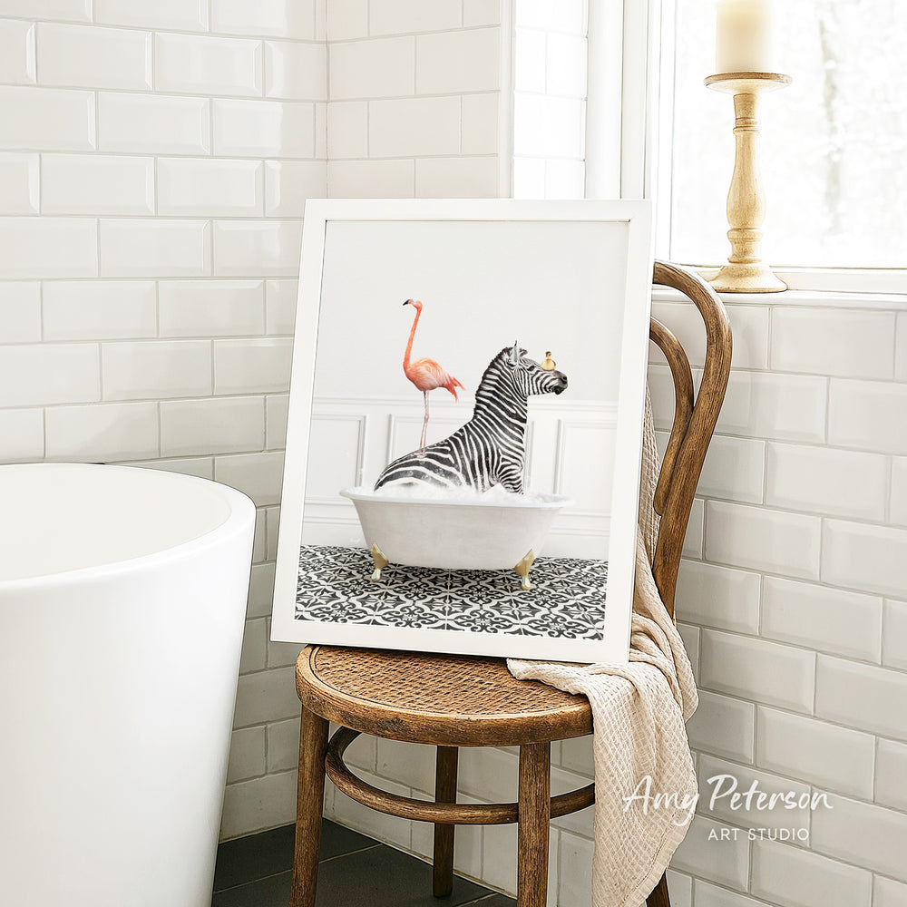 A framed image of a zebra sitting in a bathtub, with a pink flamingo standing on its back, is displayed on a wooden chair in a bathroom setting.