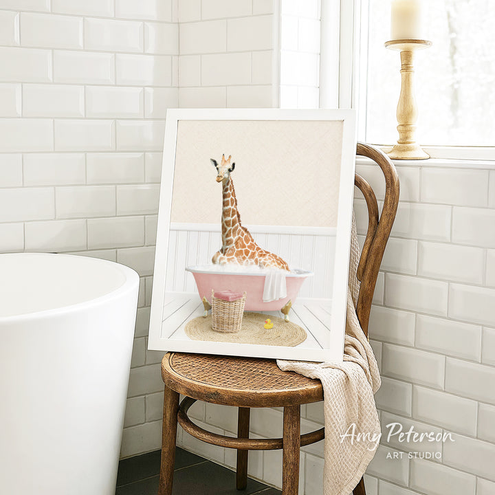 A framed image of a giraffe sitting in a pink bathtub, surrounded by a basket of toys, is displayed on a wooden chair in a bathroom setting.