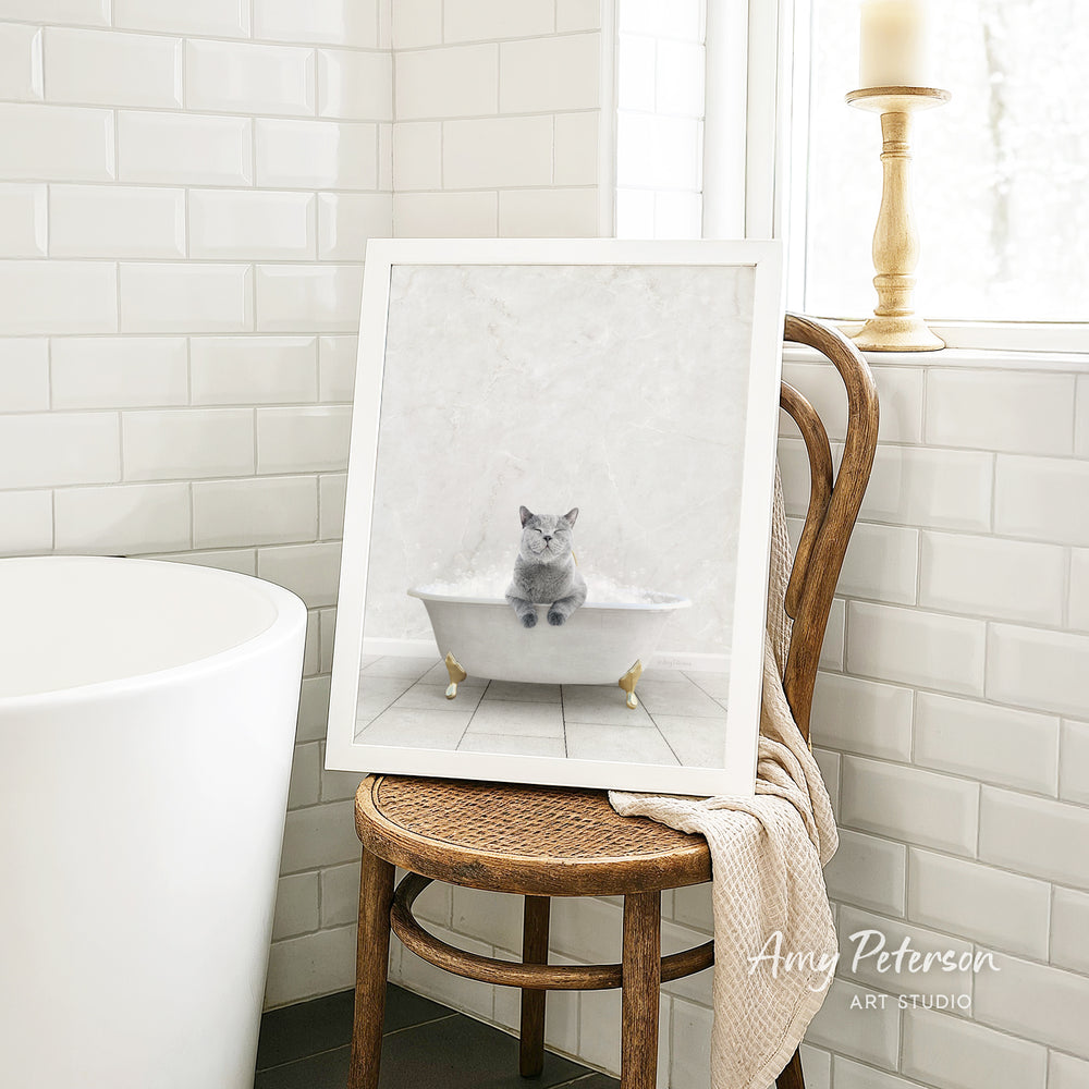 A framed image of a cat sitting in a bathtub, with a towel draped over the back of a wooden chair in a bathroom setting.