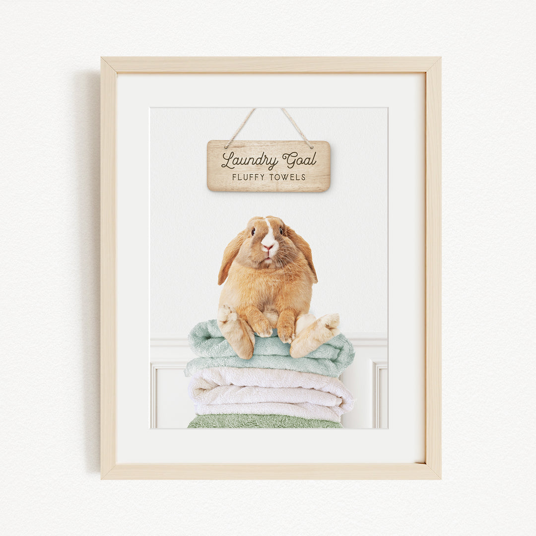 A framed image of a rabbit sitting on a stack of folded towels, with a wooden sign above that says "Laundry goal Fluffy towels".