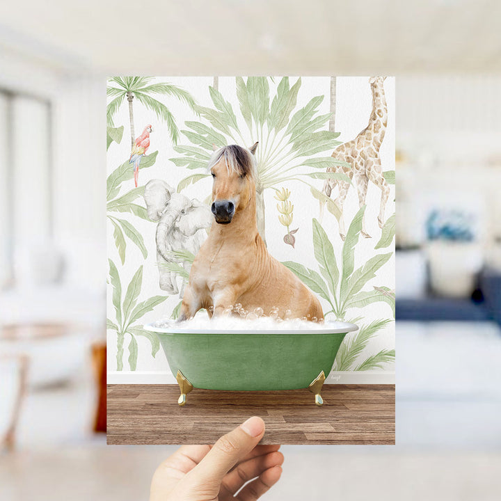 A hand holds up a framed image depicting a horse sitting in a bathtub surrounded by tropical foliage, including a giraffe and palm trees.