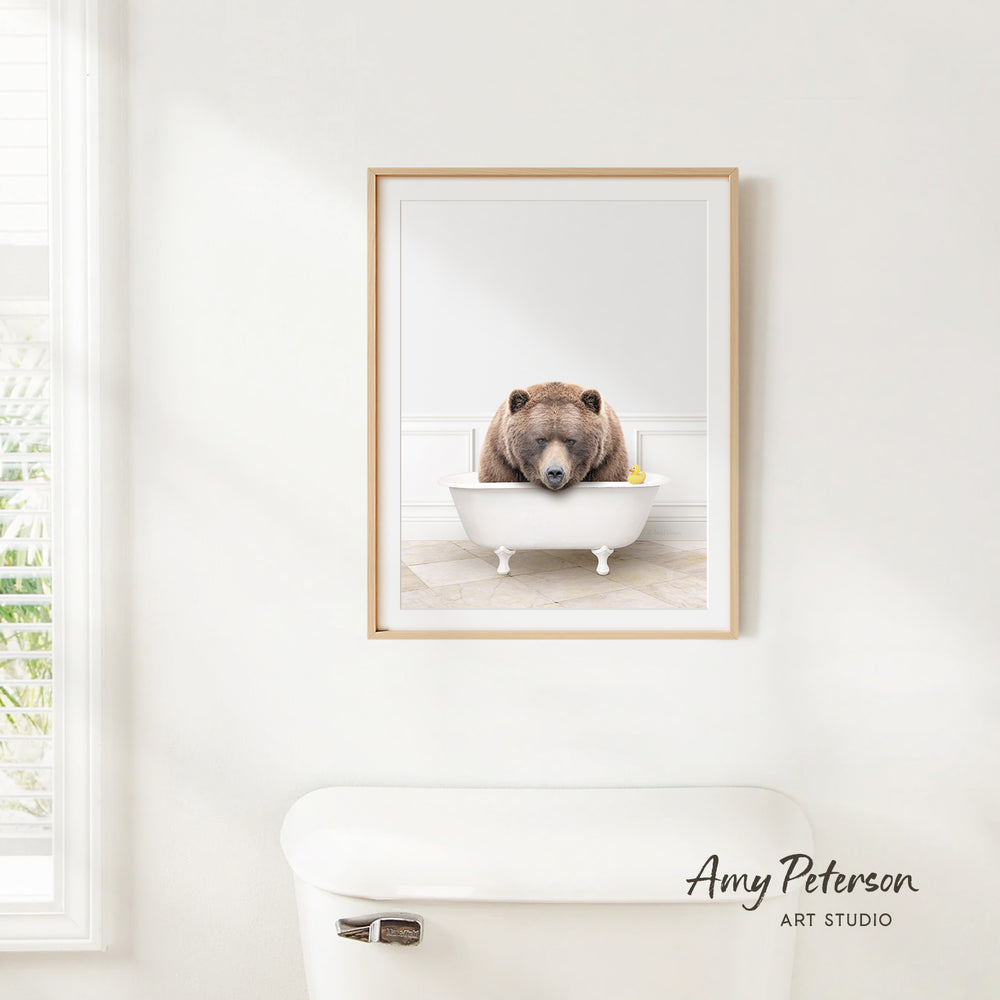 A framed image of a brown bear sitting in a white bathtub, with a yellow rubber duck floating in the water.