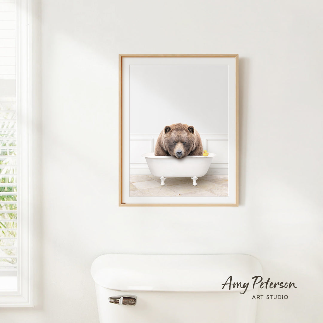 A framed image of a brown bear sitting in a white bathtub, with a yellow rubber duck floating in the water.