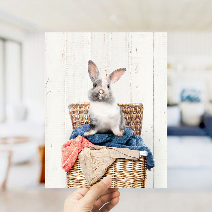 A small gray and white rabbit sitting in a wicker basket filled with clothes and blankets.