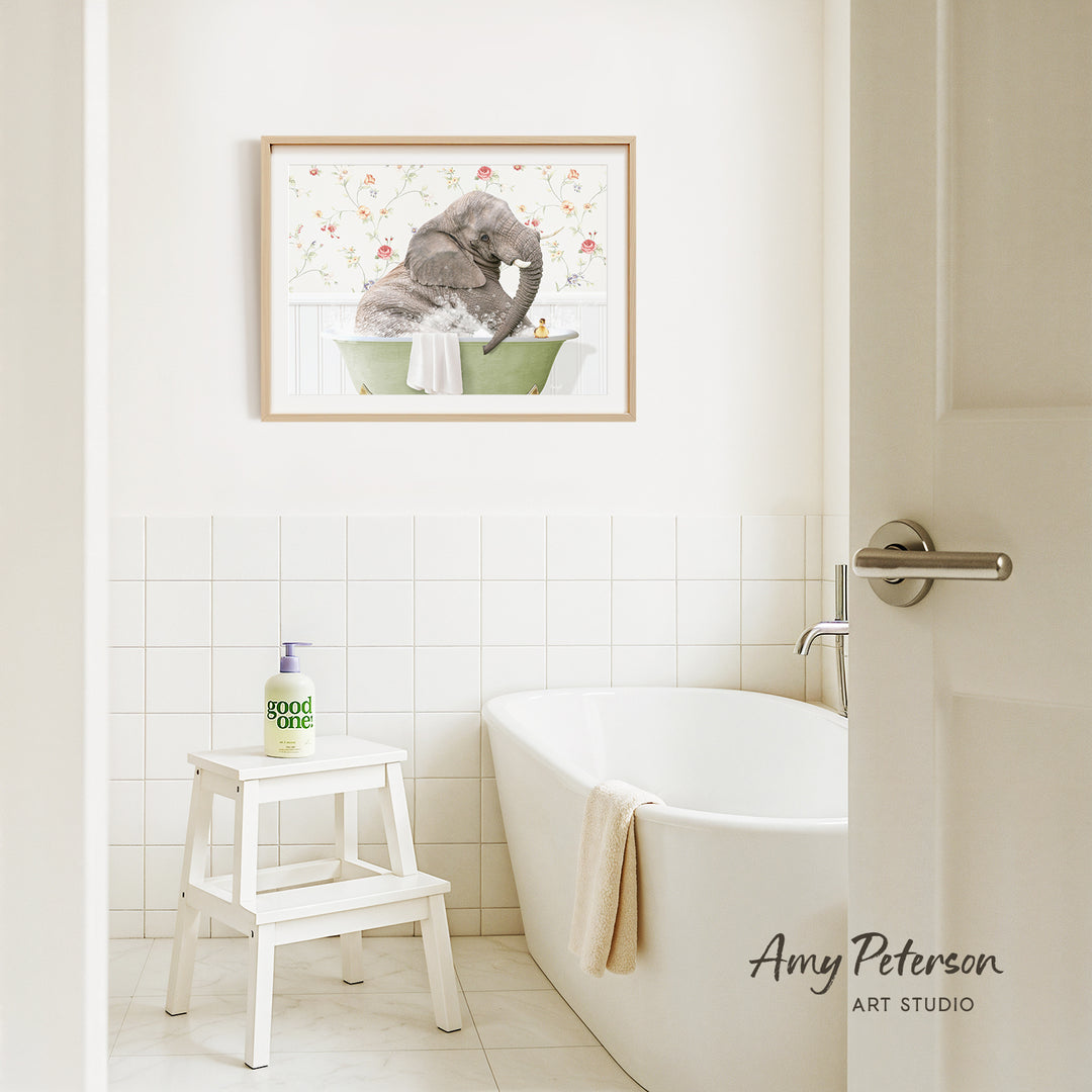 A framed image of an elephant sitting in a bathtub is hanging on a white tiled wall above a white bathtub in a bathroom.