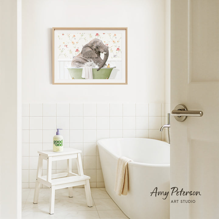 A framed image of an elephant sitting in a bathtub is hanging on a white tiled wall above a white bathtub in a bathroom.