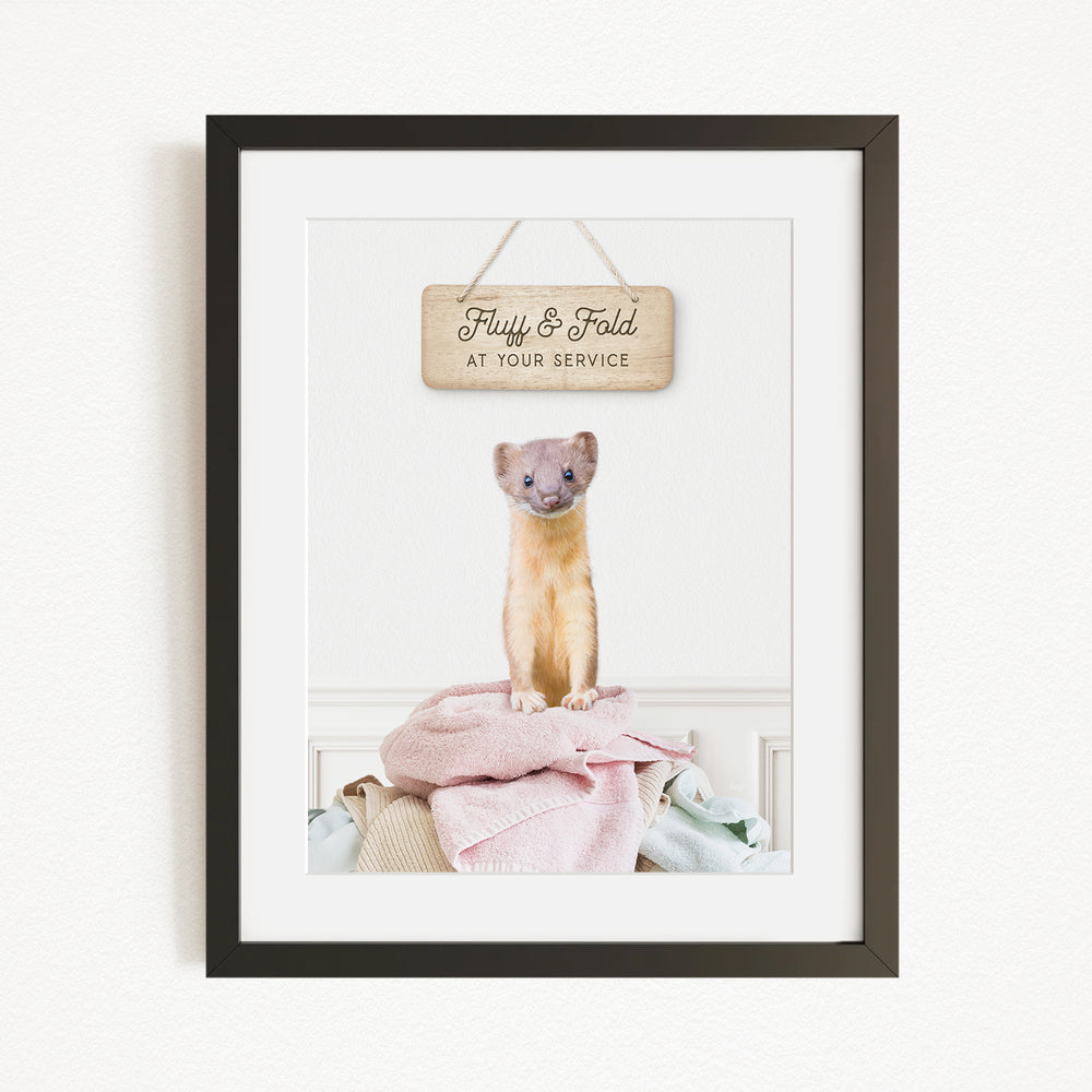 A framed image of a small, furry animal, likely a cat, sitting on a pile of clothes and towels. Above the animal, there is a wooden sign that reads "Fluffy & Fold at your service".
