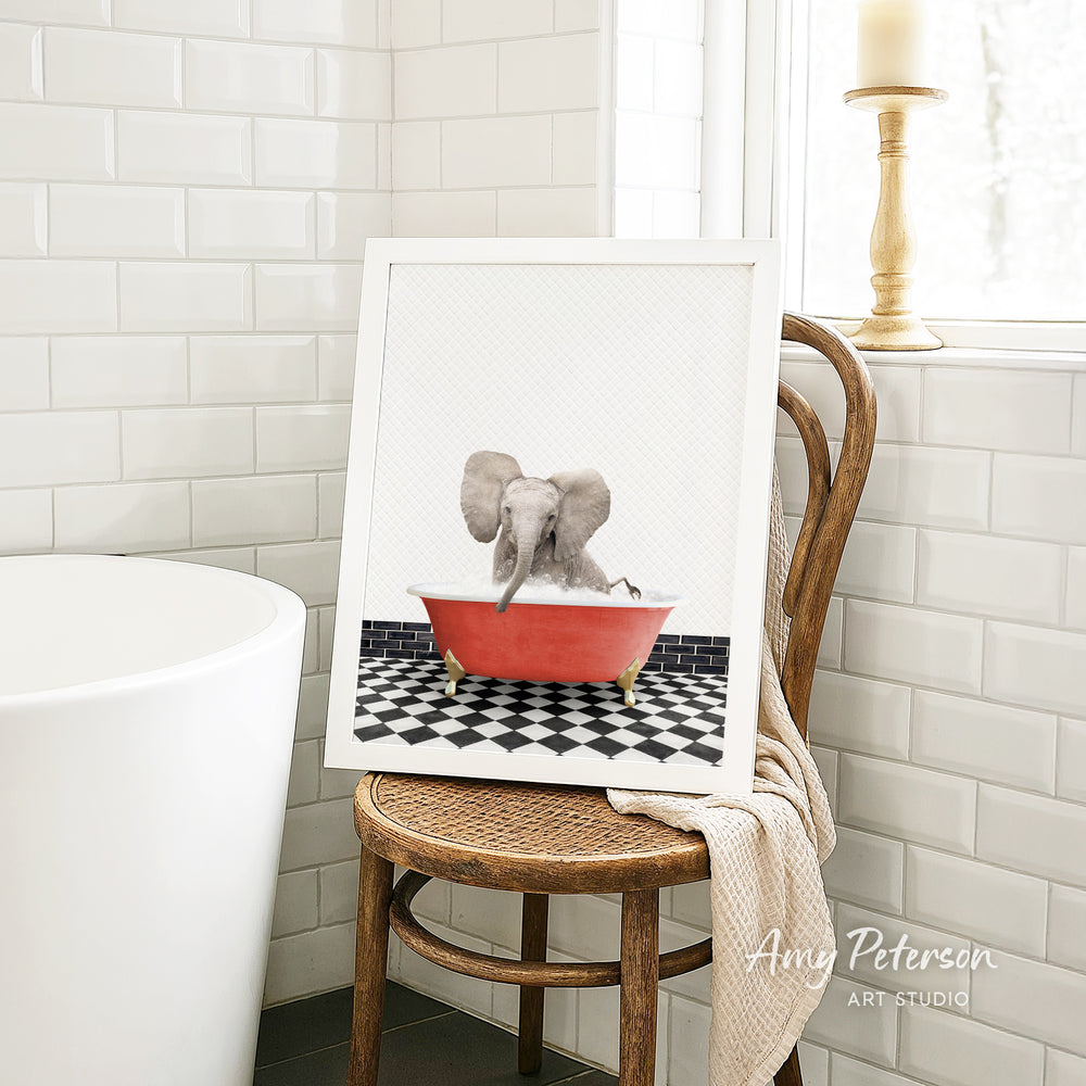A framed image of an elephant sitting in a red bathtub, surrounded by a black and white checkered floor, is displayed on a wooden chair in a bathroom setting.