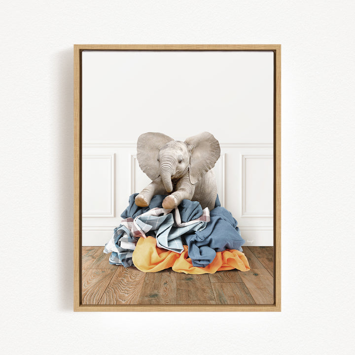A framed image of a baby elephant sitting on a pile of clothes on a wooden floor.