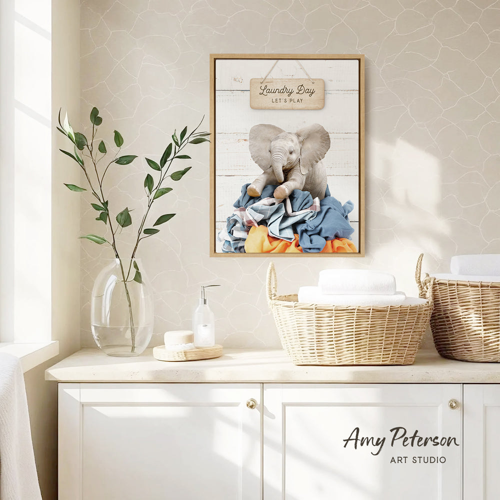 a framed poster of an elephant sitting on laundry hanging on a wall, with a wicker basket and a plant on a counter below it.