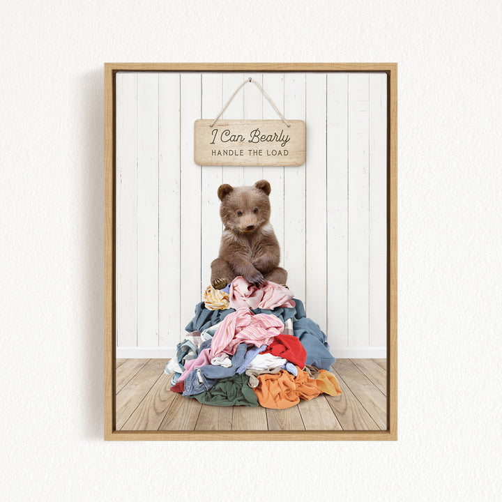 A teddy bear sitting atop a pile of clothes, with a wooden sign above that reads "I Can Bearly Handle the Load".