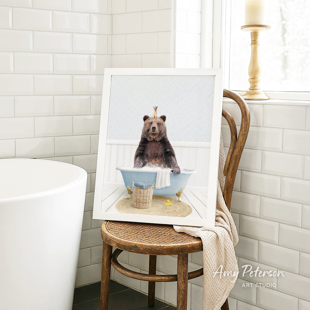 A framed image of a bear sitting in a bathtub, surrounded by a basket of bath toys, is displayed on a wooden chair in a bathroom setting.