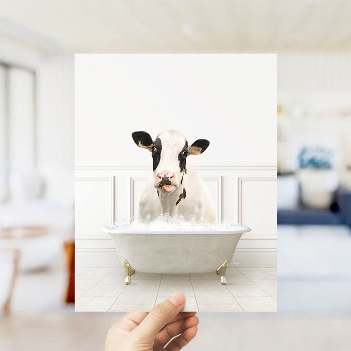 A hand holds up a framed image of a cow sitting in a bathtub filled with water, creating a whimsical and humorous scene.