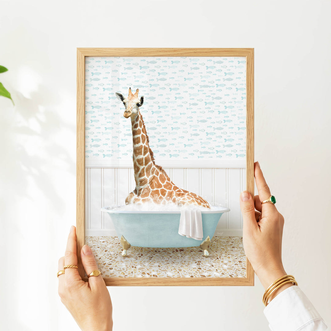 A person's hands are holding a framed picture of a giraffe sitting in a bathtub filled with water, surrounded by fish-themed wallpaper.