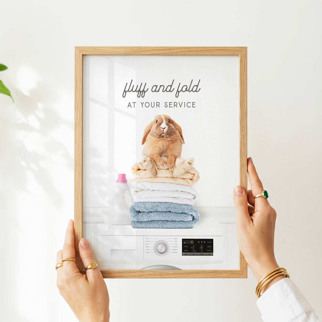 A person's hands are holding a framed picture of a rabbit sitting on top of a stack of folded towels, with a washing machine visible below.