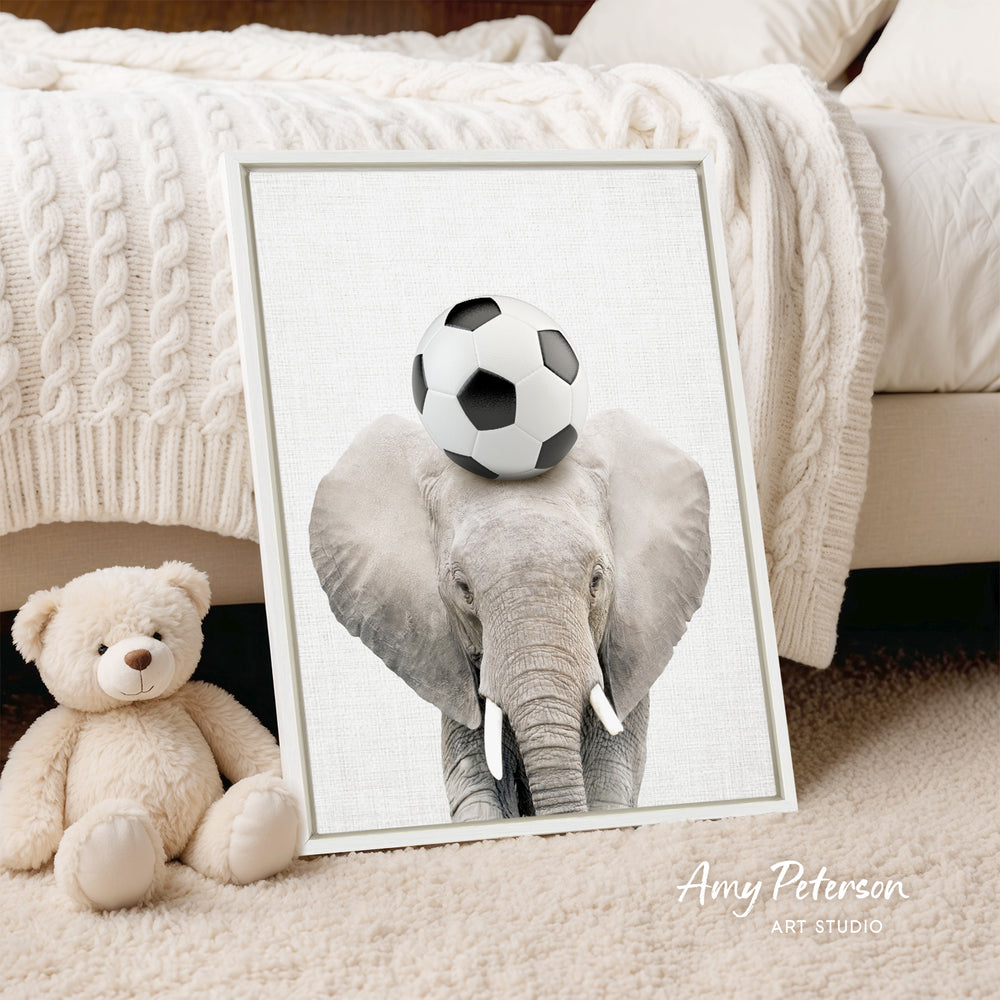 A framed image of an elephant holding a soccer ball on its head, placed on a bed next to a teddy bear.