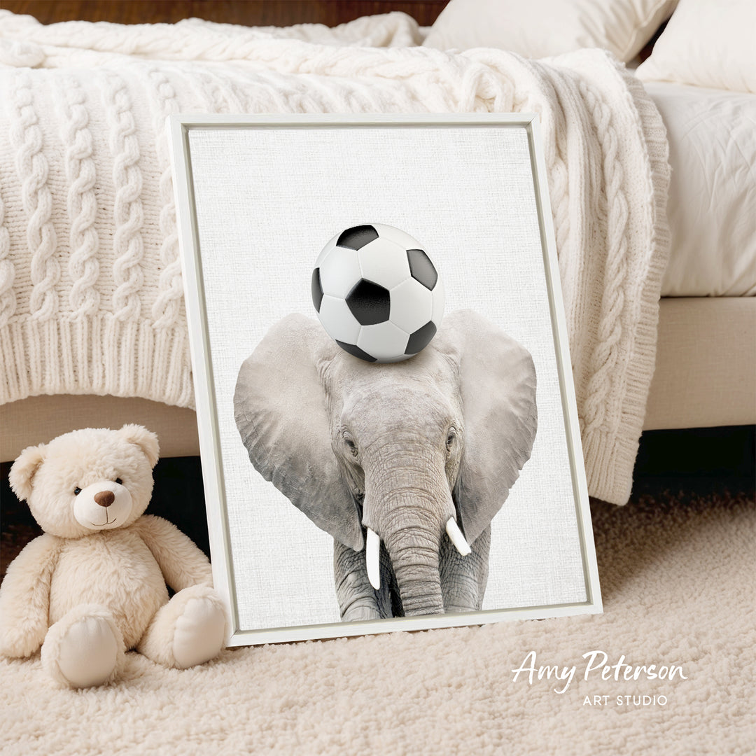A framed image of an elephant holding a soccer ball on its head, placed on a bed next to a teddy bear.