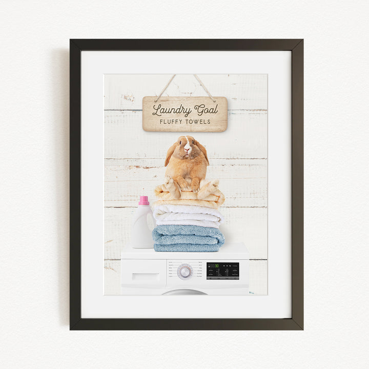 A framed image depicting a rabbit sitting on a stack of fluffy towels, with a sign above that reads "Laundry goal Fluffy towels".