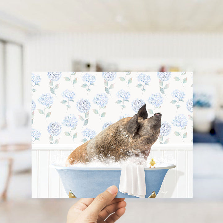 A hand holding a framed image of a pig sitting in a bathtub filled with bubbles, surrounded by a floral pattern.