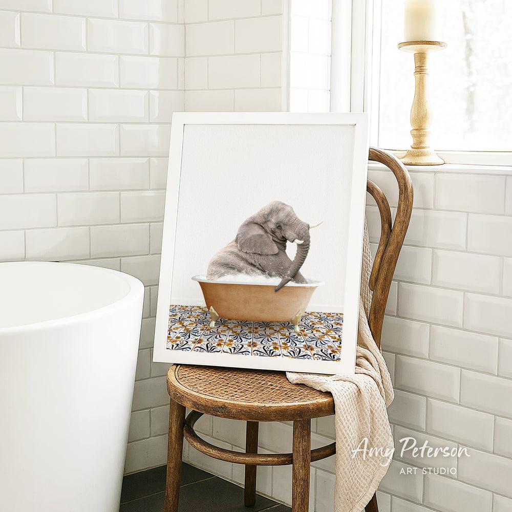 A framed image of a gray elephant sitting in a bathtub, surrounded by a patterned design, is displayed on a wooden chair in a bathroom setting.