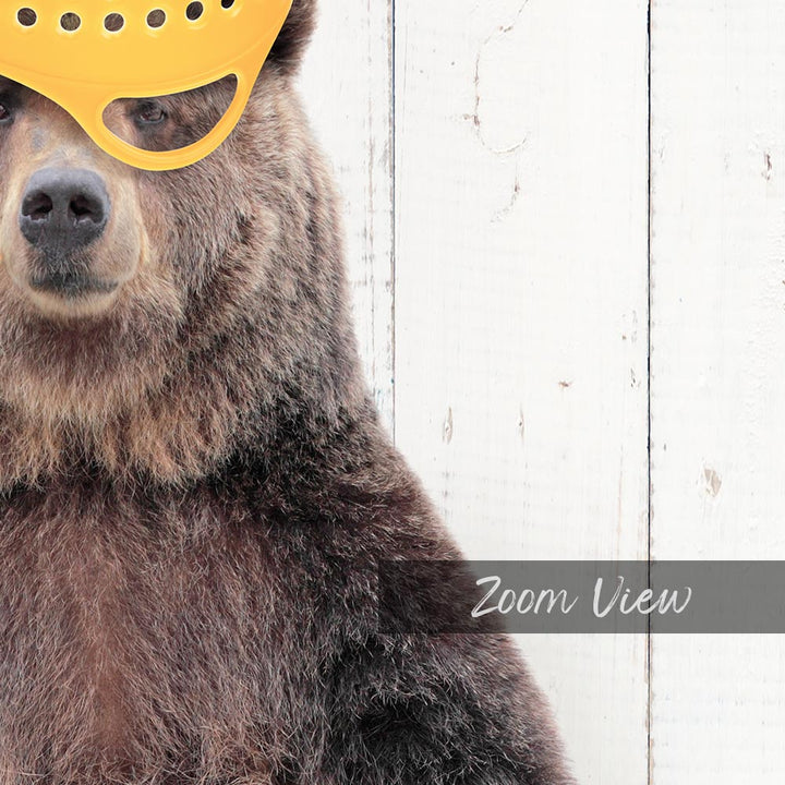 A close-up of a bear wearing a yellow plastic mask, with a white wooden background.