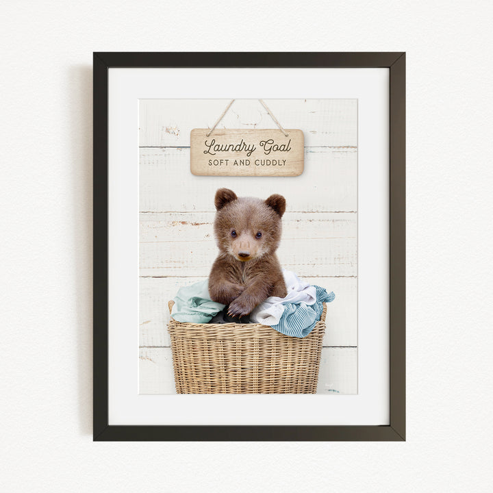 A framed image of a baby bear sitting in a wicker basket filled with clothes, with a wooden sign above that says "Laundry Goal Soft and Cuddly".