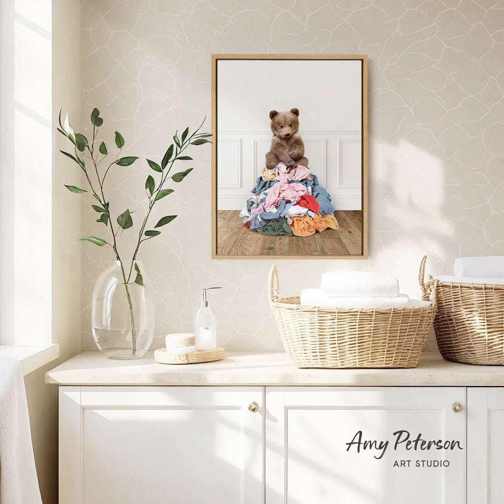 a framed photograph of a teddy bear sitting on a pile of clothes, hanging on a wall above a white cabinet.