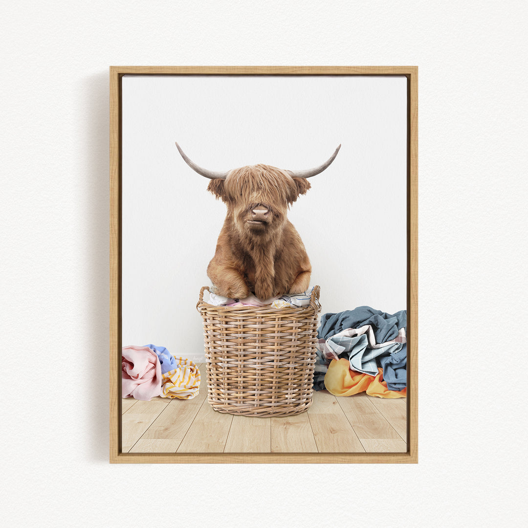 A Highland cow with long horns is sitting in a wicker basket filled with clothes, creating a whimsical and playful scene.