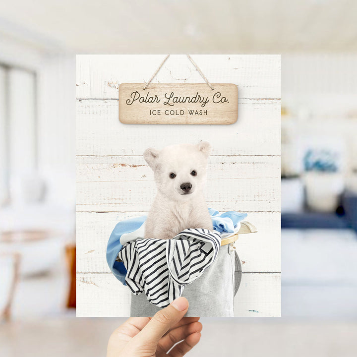 A hand holding a framed photograph of a white polar bear sitting in a laundry basket, with a wooden sign above it that reads "Polar Laundry Co. ICE COLD WASH".