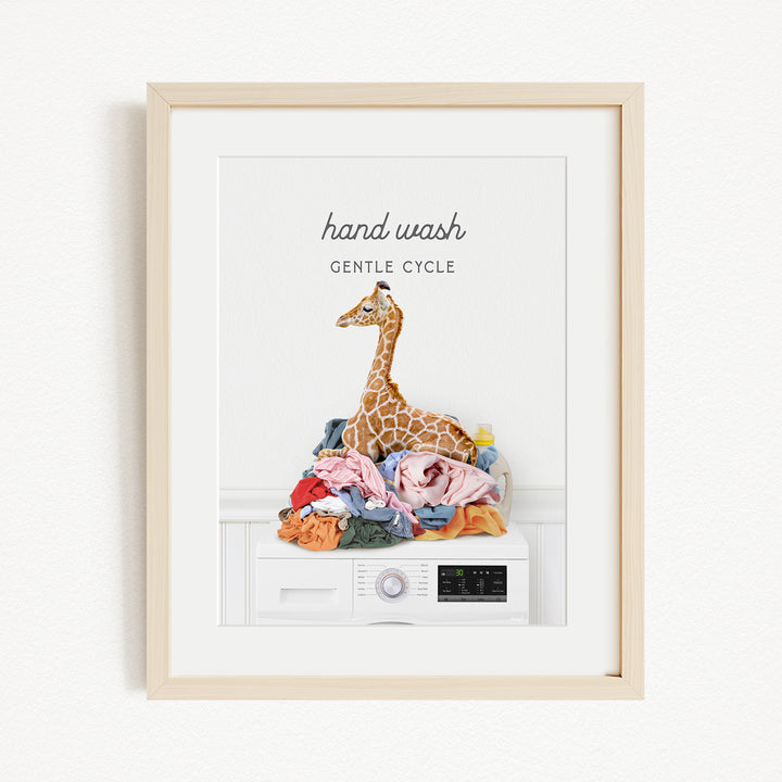 A framed image depicting a giraffe sitting atop a pile of laundry, with the text "hand wash gentle cycle" written above it.