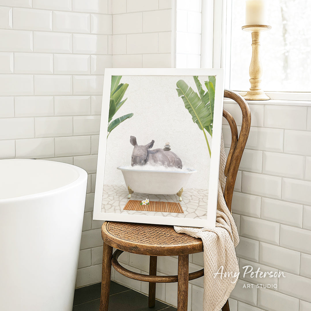 A framed painting of a rhinoceros sitting in a bathtub, surrounded by tropical plants, is displayed on a wooden chair in a bathroom setting.
