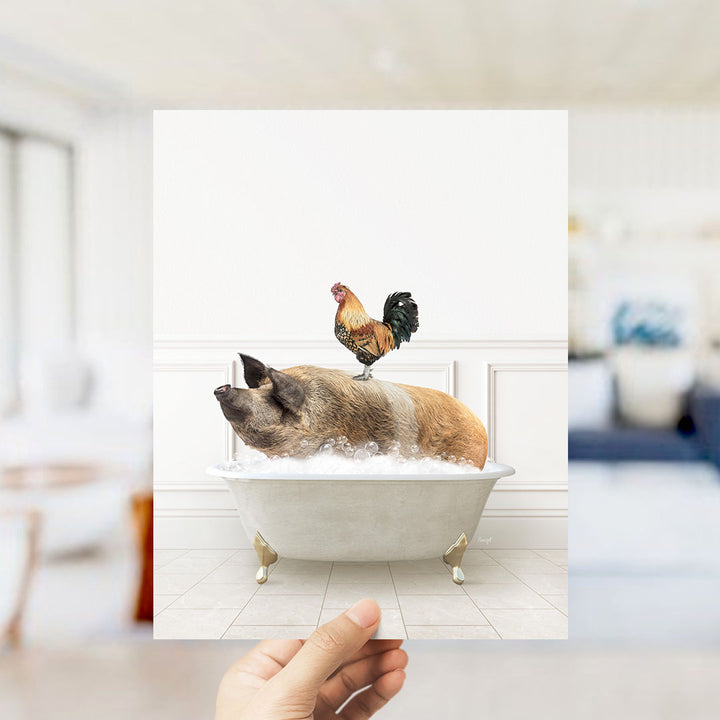 A hand holds up a framed image depicting a pig sitting in a bathtub, with a rooster perched on its back.