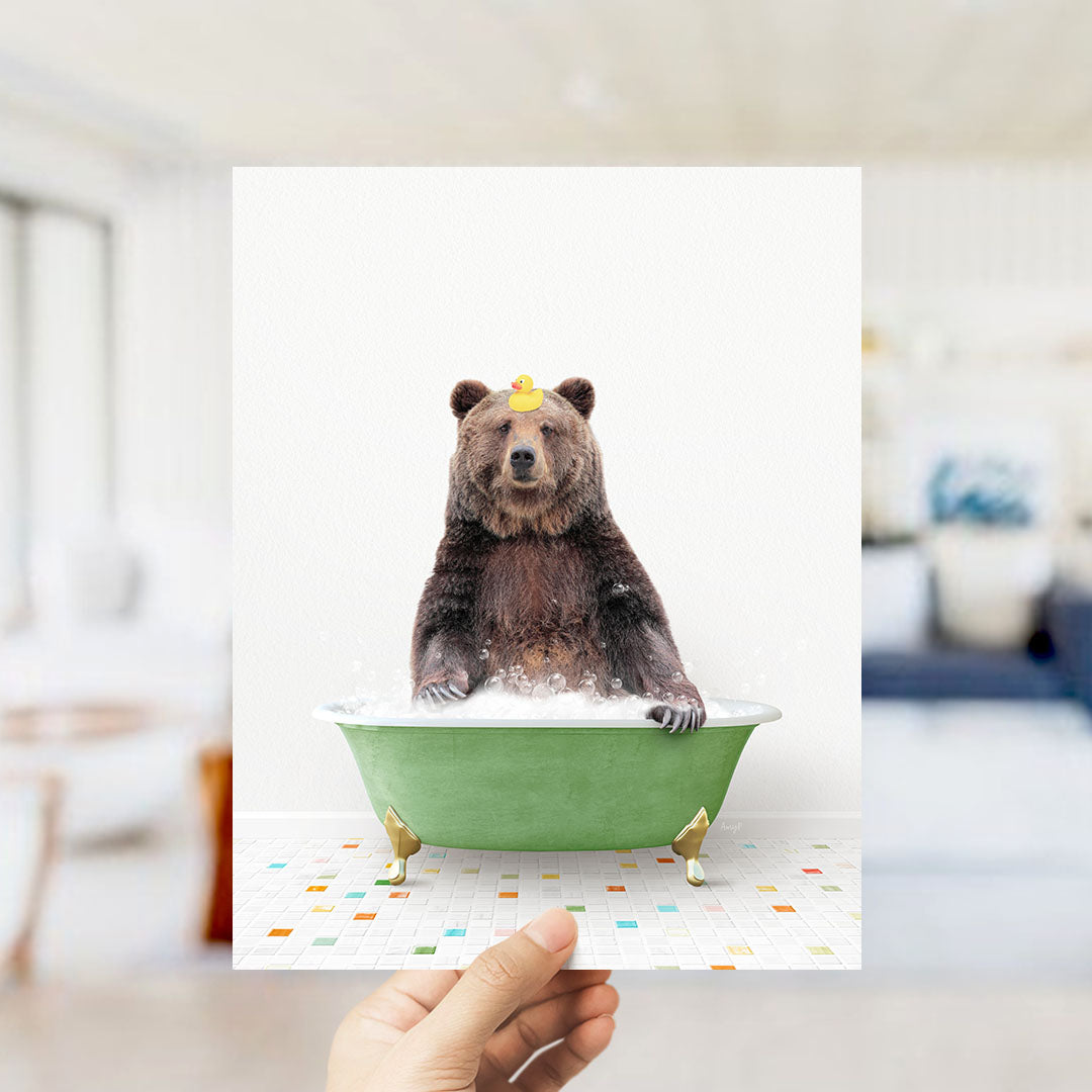 A hand holds up a framed image of a brown bear sitting in a green bathtub filled with bubbles, wearing a yellow rubber duck.