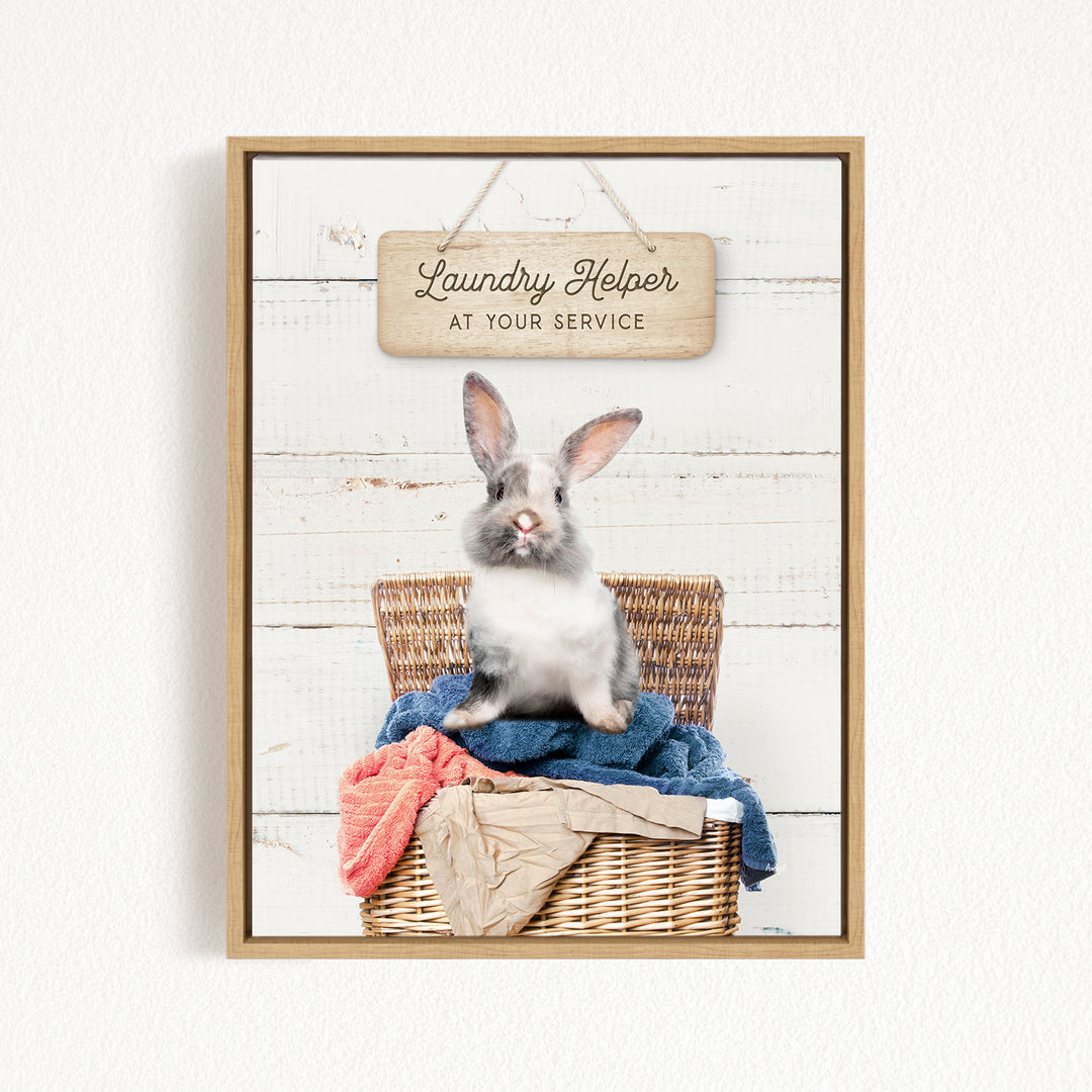 A gray and white rabbit sitting in a wicker laundry basket filled with clothes, with a wooden sign above that reads "Laundry Helper at your service".