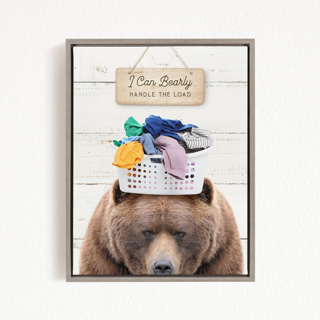 A brown bear sits in front of a laundry basket filled with clothes, with a wooden sign above that reads "I Can Bearly Handle the Load".