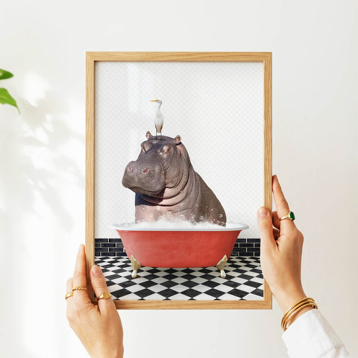 A person's hands are holding a framed picture of a hippopotamus sitting in a bathtub filled with water, with a bird perched on its head.