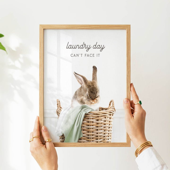 A person's hands are holding a picture frame with a photograph of a rabbit sitting in a wicker basket, with the text "laundry day can't face it" written above the image.