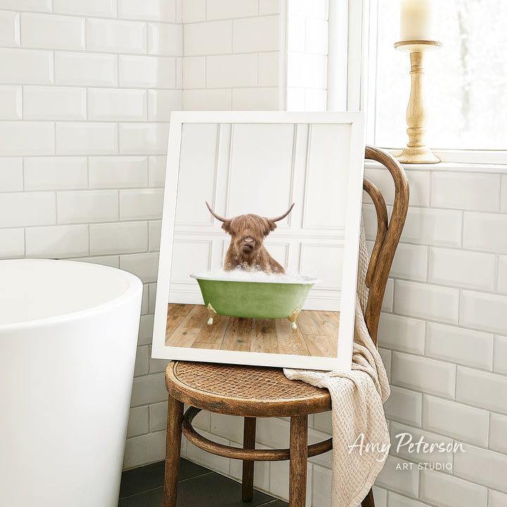 A framed image of a Highland cow sitting in a green bathtub, with a towel draped over the back of a wooden chair in a bathroom setting.
