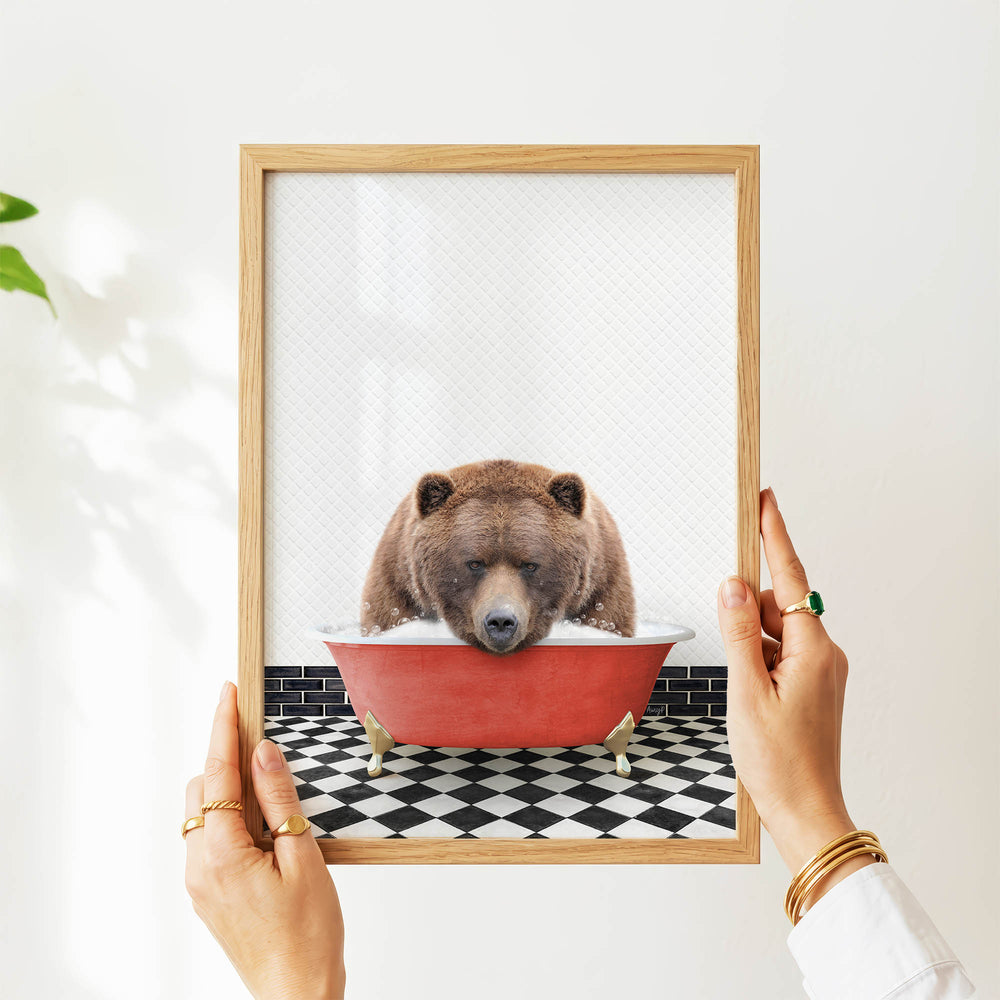 A person's hands are holding a framed image of a brown bear sitting in a red bathtub, with a black and white checkered floor visible beneath the tub.