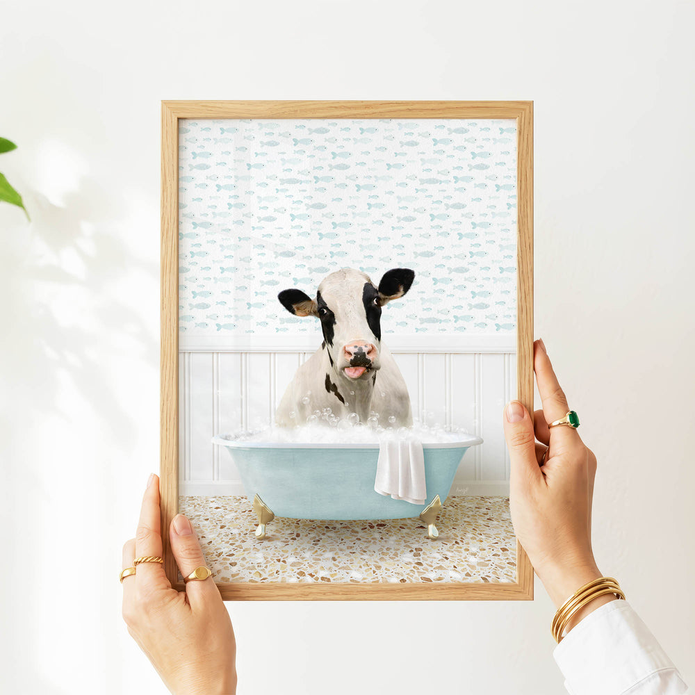 A person's hands are holding a framed picture of a cow sitting in a bathtub filled with bubbles, with a patterned wall behind it.