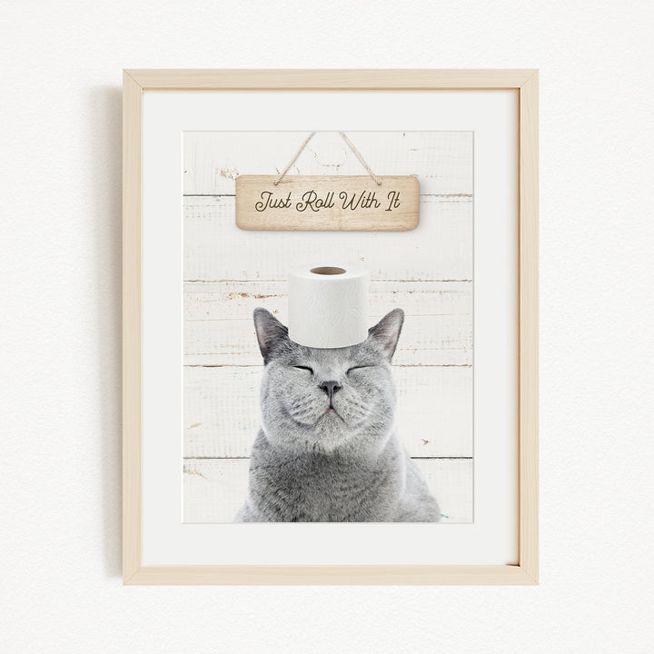 A framed image of a gray cat with its eyes closed, wearing a roll of toilet paper on its head, and a wooden sign above it that says "Just Roll With It".