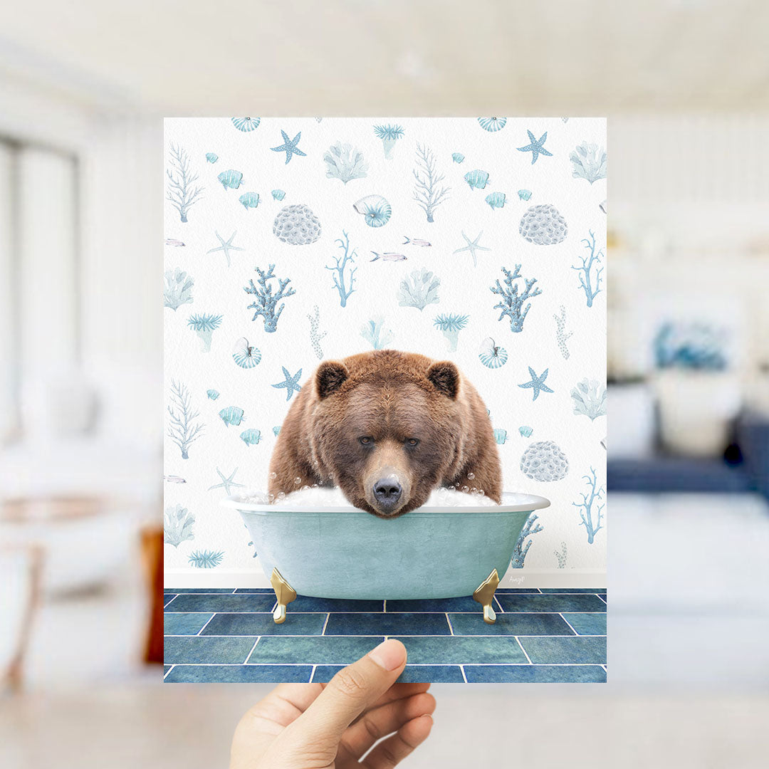 A hand is holding up a print featuring a brown bear sitting in a bathtub filled with bubbles, surrounded by a pattern of seashells, starfish, and other marine life.