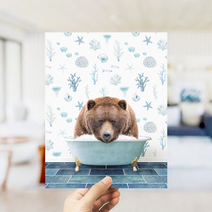 A hand is holding up a print featuring a brown bear sitting in a bathtub filled with bubbles, surrounded by a pattern of seashells, starfish, and other marine life.