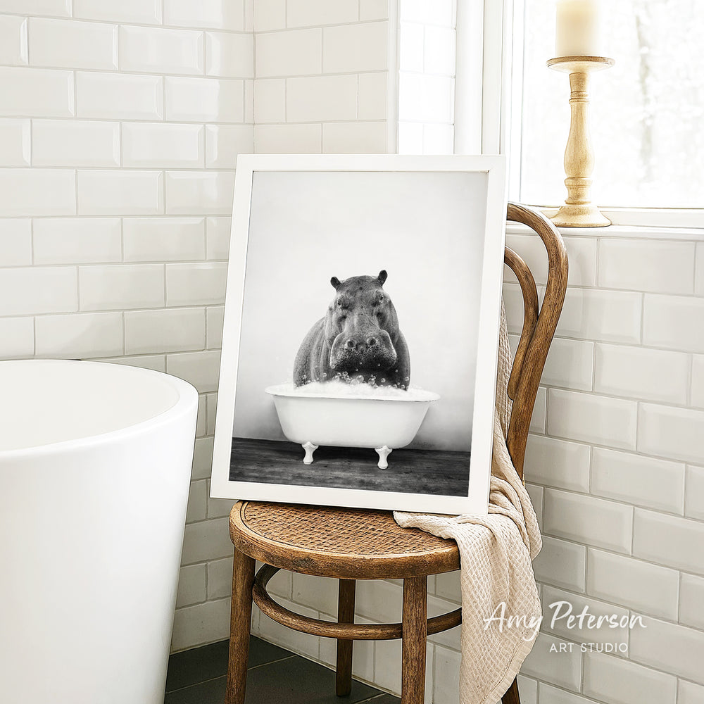 A framed black and white photograph of a hippopotamus sitting in a bathtub is displayed on a wooden chair in a bathroom setting.