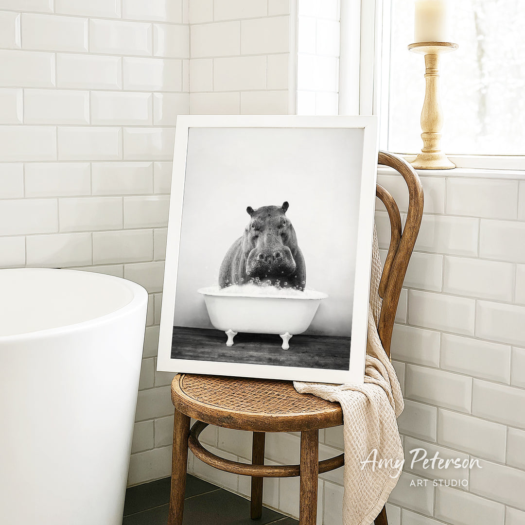 A framed black and white photograph of a hippopotamus sitting in a bathtub is displayed on a wooden chair in a bathroom setting.