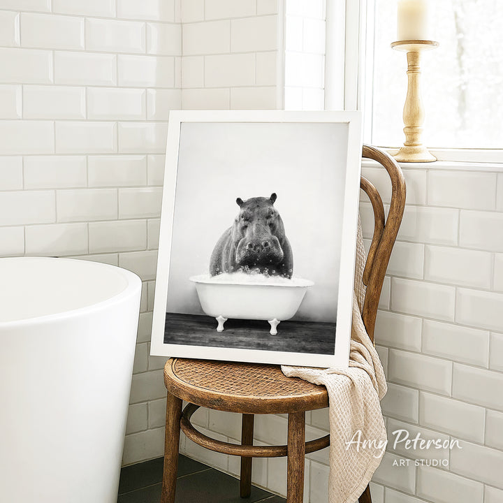 A framed black and white photograph of a hippopotamus sitting in a bathtub is displayed on a wooden chair in a bathroom setting.