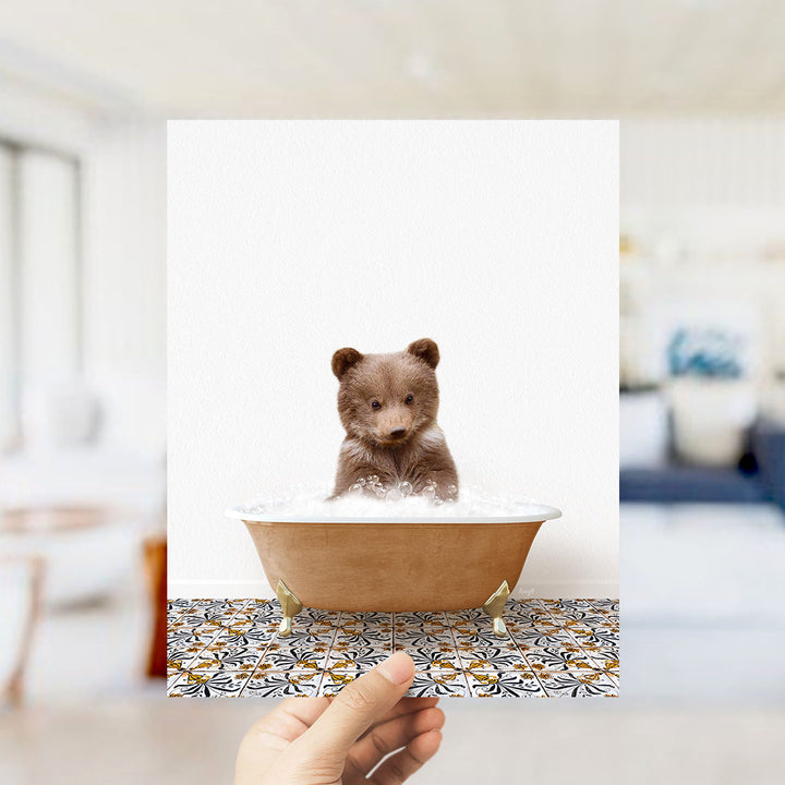 A hand holds up a framed image of a small brown bear sitting in a bathtub filled with bubbles, surrounded by a patterned tile floor.