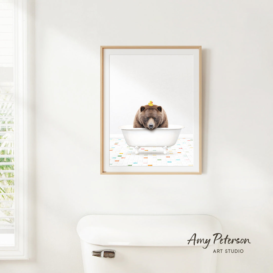A framed image of a brown bear sitting in a white bathtub, with a yellow object on its head, is hanging on a white wall.