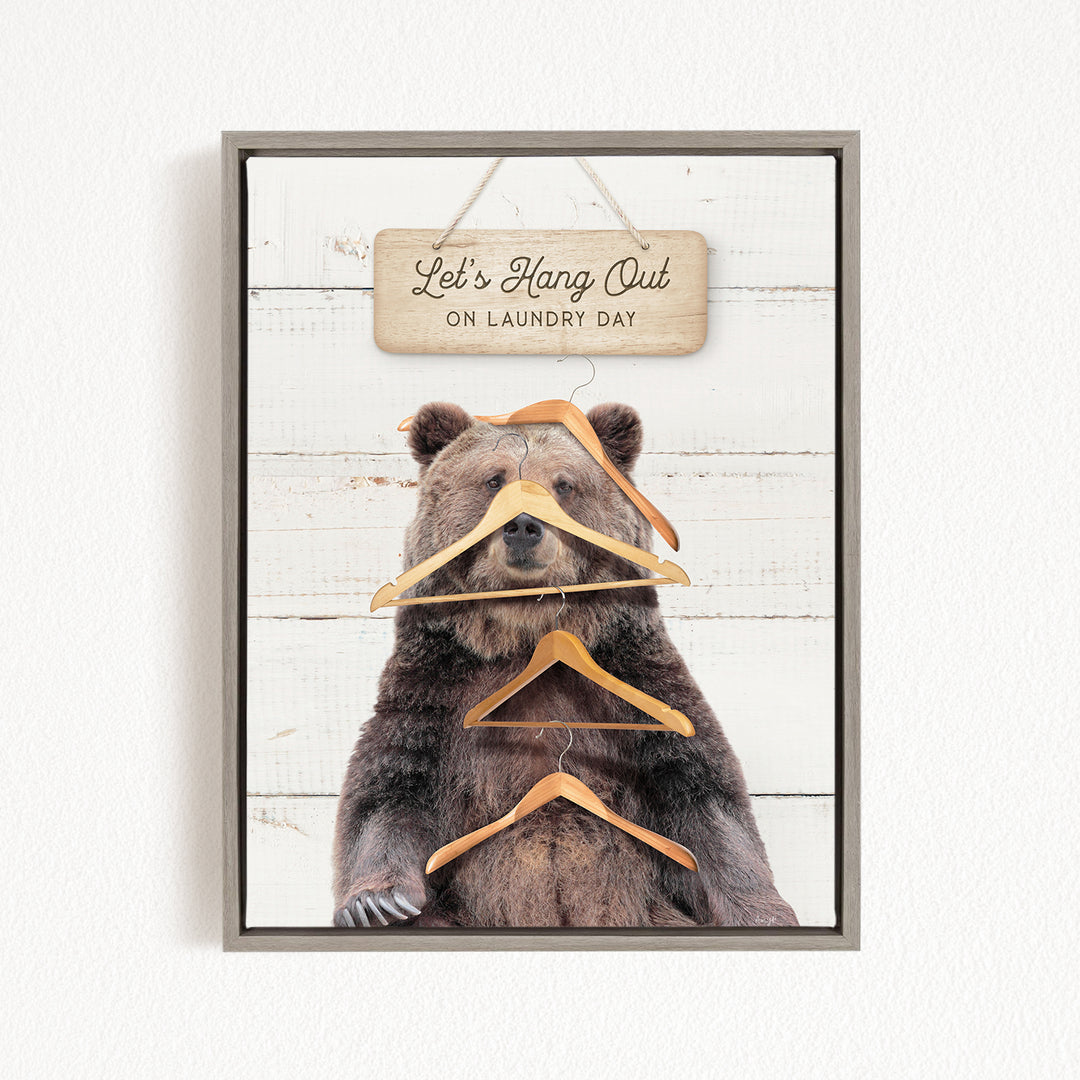 A framed image of a bear holding multiple wooden hangers, with a sign above that says "Let's Hang Out on Laundry Day".